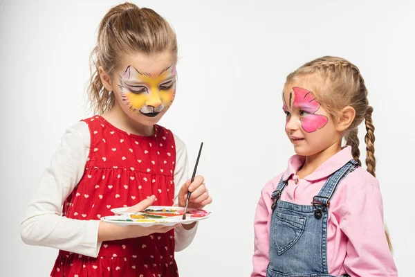 Mignon enfant avec muselière chat peinture sur le visage tenant palette et pinceau ami proche avec masque papillon peint isolé sur blanc — Photo de stock