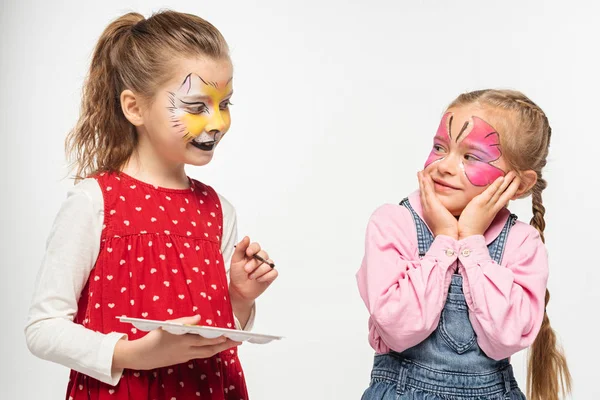 Niedliches Kind mit Katzenschnauze Malerei auf Gesicht halten Palette und Pinsel in der Nähe amüsiert Freund mit gemalten Schmetterlingsmaske isoliert auf weiß — Stockfoto