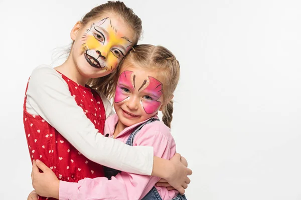 Niedliche Kinder mit Katzenschnauze und Schmetterlingsmalereien auf Gesichtern, die sich umarmen, während sie die Kamera isoliert auf Weiß betrachten — Stockfoto