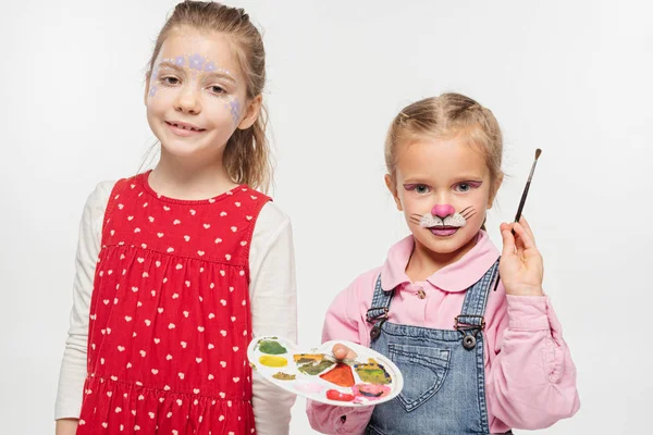 Entzückendes Kind mit Katzenschnauze Malerei des Gesichts hält Palette und Pinsel in der Nähe Freund mit gemalten floralen Maske isoliert auf weiß — Stockfoto