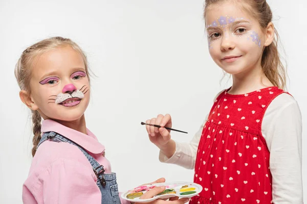 Crianças sorridentes com pinturas em rostos segurando paleta e pincel enquanto olha para a câmera isolada no branco — Fotografia de Stock