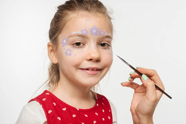 Vue recadrée de l'artiste tenant pinceau près mignon enfant avec peinture florale sur le visage isolé sur blanc — Photo de stock