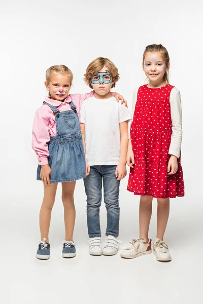 Full length view of cheerful kids and offended boy with colorful face paintings looking at camera on white background — Stock Photo
