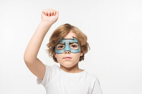 Serio ragazzo con maschera da supereroe dipinta sul viso in piedi con pugno in rilievo mentre guarda la fotocamera isolata su bianco — Foto stock