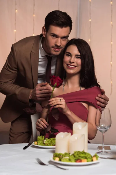 Hombre guapo abrazando feliz novia sosteniendo rosa roja mientras mira a la cámara - foto de stock
