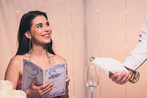 Vista recortada de camarero vertiendo vino blanco cerca de la chica feliz celebración de menú — Stock Photo