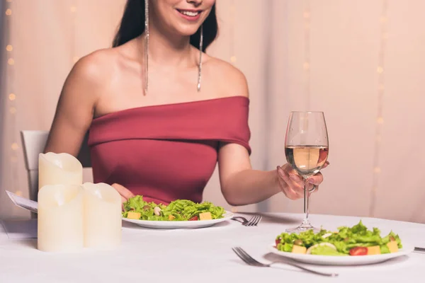 Vista recortada de chica elegante sosteniendo vaso de vino blanco mientras se sienta en la mesa servida - foto de stock