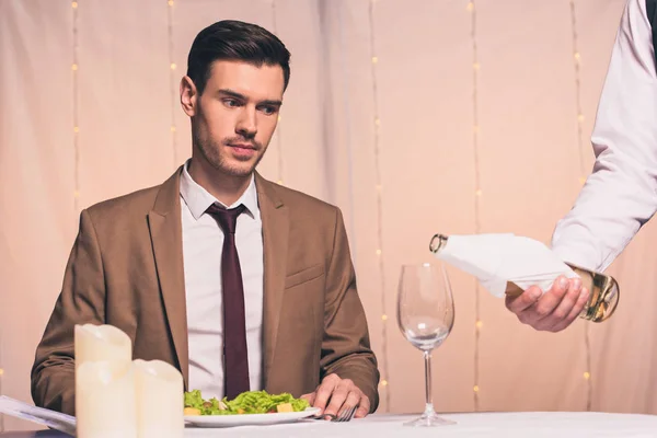 Raccolto di cameriere versando vino bianco in vetro vicino bello, uomo elegante seduto nel ristorante — Foto stock