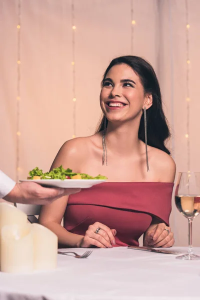 Vista recortada del camarero sosteniendo plato con ensalada fresca cerca alegre, chica elegante - foto de stock