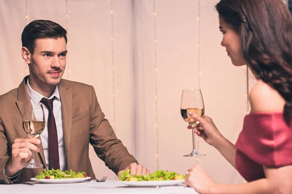 Hombre y mujer felices y elegantes sentados en la mesa servida y sosteniendo un vaso de vino blanco - foto de stock