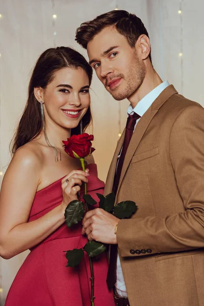 Feliz, elegante pareja mirando a la cámara mientras sostiene rosa roja en el restaurante - foto de stock