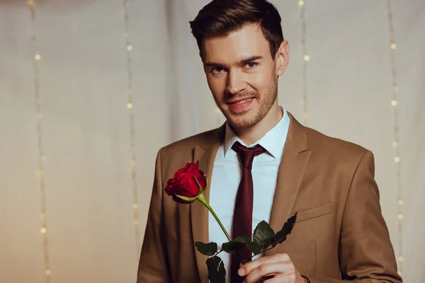 Guapo, elegante hombre sosteniendo rosa roja mientras sonríe a la cámara en el restaurante — Stock Photo