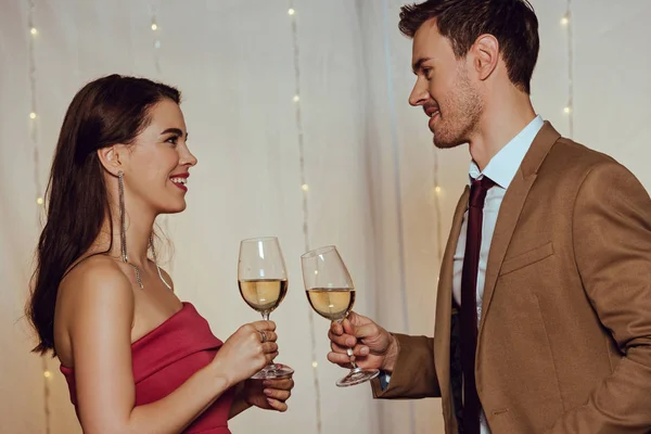 Vista lateral de pareja feliz y elegante mirándose mientras sostiene vasos de vino blanco - foto de stock