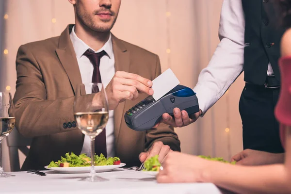 Vista recortada de un hombre elegante que paga con tarjeta de crédito con terminal mientras está sentado en el restaurante cerca de la novia - foto de stock