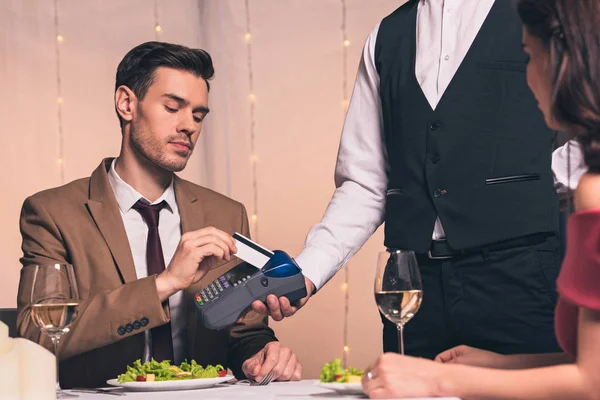 Guapo, elegante hombre que paga con tarjeta de crédito con terminal mientras está sentado en el restaurante cerca de la novia - foto de stock