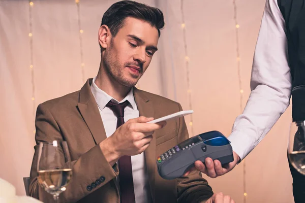 Vista parcial del camarero con terminal de pago cerca del elegante hombre que sostiene la tarjeta de crédito mientras está sentado en el restaurante - foto de stock