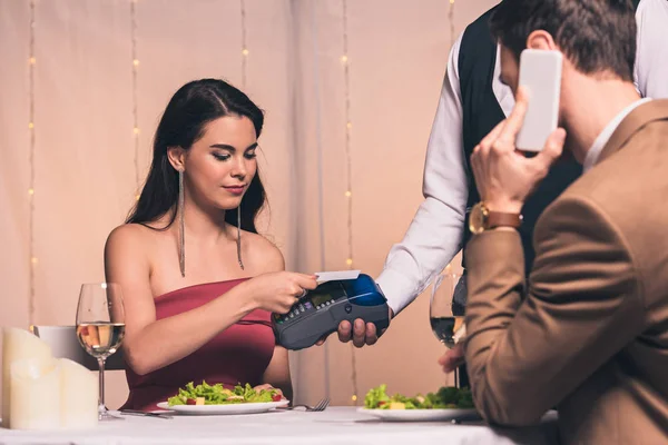 Chica elegante que paga con tarjeta de crédito con terminal en el restaurante mientras que el novio habla en el teléfono inteligente - foto de stock