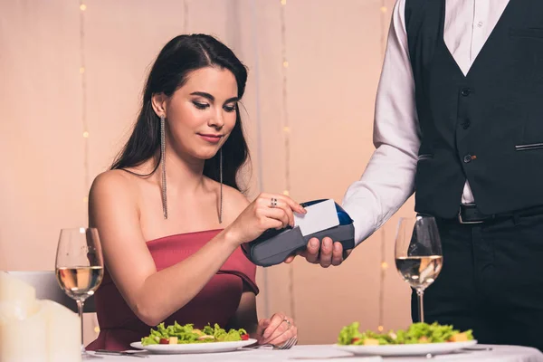 Vista recortada de camarero sosteniendo terminal de pago cerca hermosa, chica elegante sentada a la mesa servida - foto de stock