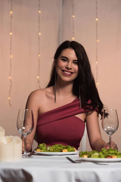 Felice, ragazza elegante seduta al tavolo servito nel ristorante e sorridente alla macchina fotografica — Foto stock