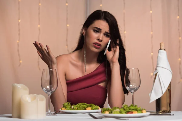 Ofendido, chica elegante sentada en la mesa servida con el brazo abierto mientras habla en el teléfono inteligente - foto de stock