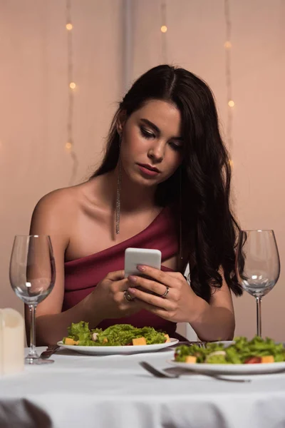 Chica elegante, decepcionado sentado en la mesa servida y el uso de teléfono inteligente en el restaurante - foto de stock