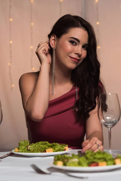 Усміхнена, елегантна дівчина торкається волосся, сидячи за поданим столом в ресторані — стокове фото