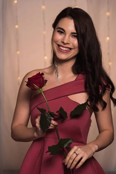 Счастливая, элегантная девушка, держащая красную розу и улыбающаяся камере в ресторане — стоковое фото