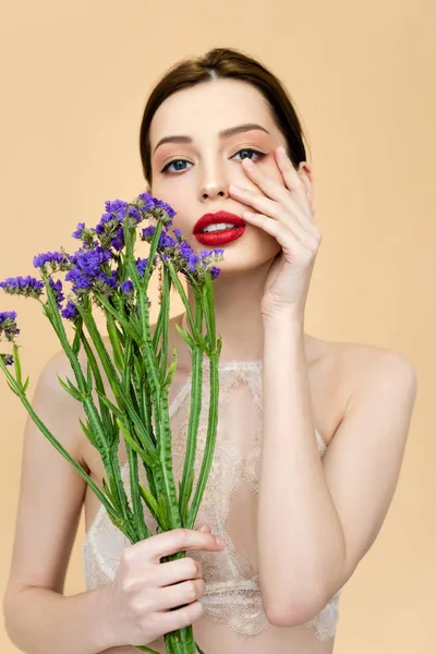 Belle femme regardant caméra et toucher le visage tout en tenant des fleurs de limonium isolé sur beige — Photo de stock