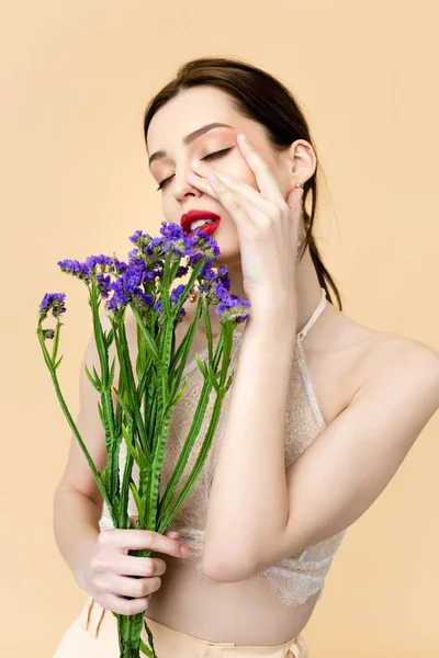 Beautiful woman with closed eyes touching face and holding purple limonium flowers isolated on beige — Stock Photo