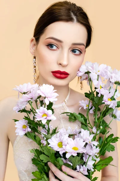 Mujer joven sosteniendo flores de crisantemo en flor y mirando hacia otro lado aislado en beige - foto de stock
