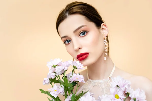 Bonita mujer joven mirando a la cámara cerca de flores de crisantemo florecientes aisladas en beige - foto de stock