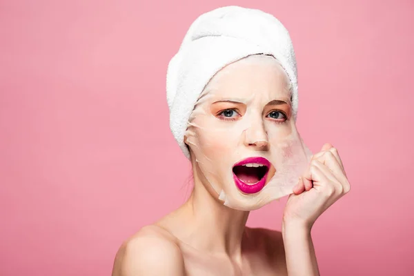 Überraschte junge Frau berührt Gesichtsmaske isoliert auf rosa — Stockfoto