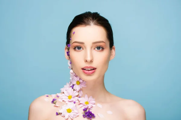 Nackte junge Frau mit Blumen, die isoliert auf blauem Grund in die Kamera blickt — Stockfoto