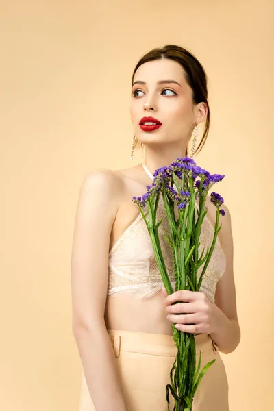 Jeune femme séduisante tenant des fleurs de limonium et regardant loin isolé sur beige — Photo de stock