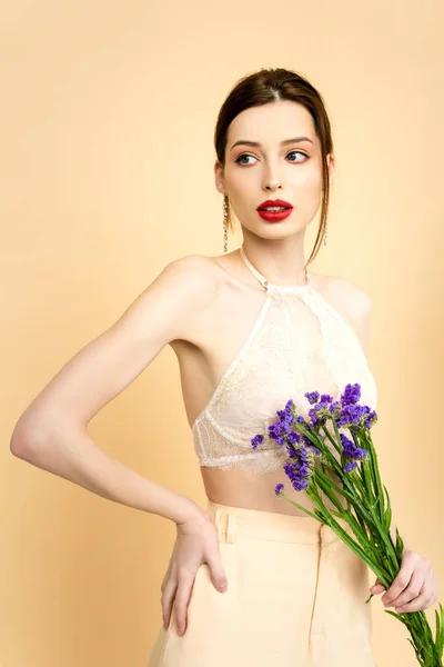 Giovane donna attraente che tiene fiori di limonio viola e in piedi con mano sul fianco isolato su beige — Foto stock