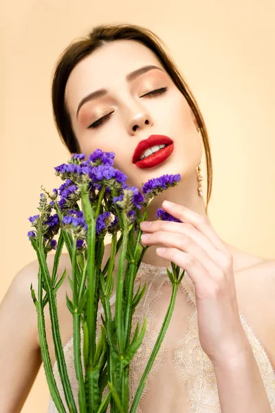Bonita mujer con los ojos cerrados sosteniendo flores de limonio púrpura aislado en beige - foto de stock