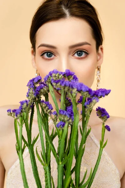 Giovane donna attraente che copre il viso con fiori di limonio viola isolati sul beige — Foto stock