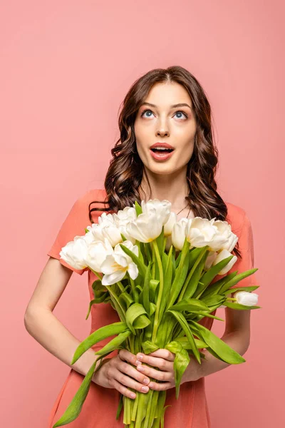 Chica sorprendida mirando hacia arriba mientras sostiene ramo de tulipanes blancos aislados en rosa - foto de stock