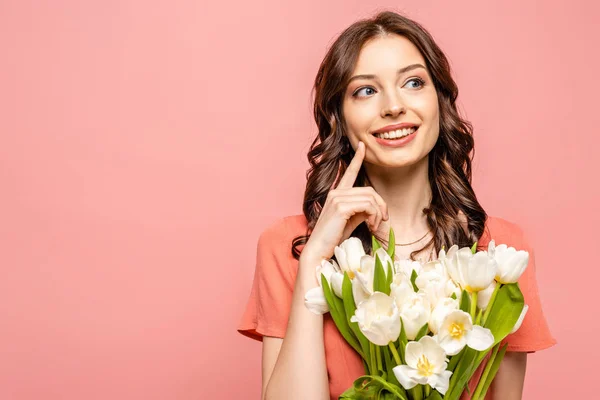 Happy girl looking away and touching cheek while holding bouquet of white tulips isolated on pink — Stock Photo