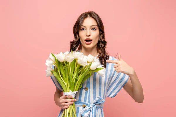 Surprised girl pointing with finger at bouquet of white tulips while looking at camera isolated on pink — Stock Photo