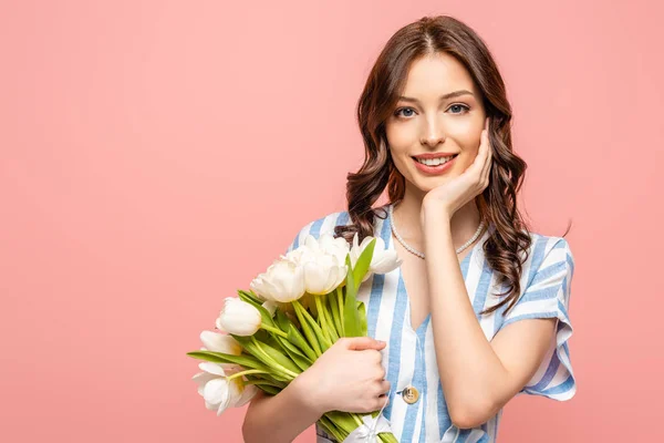 Happy girl smiling at camera while holding bouquet of white tulips and touching face isolated on pink — Stock Photo