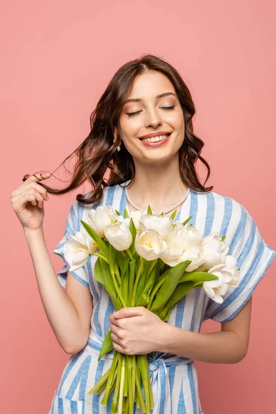 Ragazza allegra toccare i capelli mentre tiene bouquet di tulipani bianchi con gli occhi chiusi isolati su rosa — Foto stock