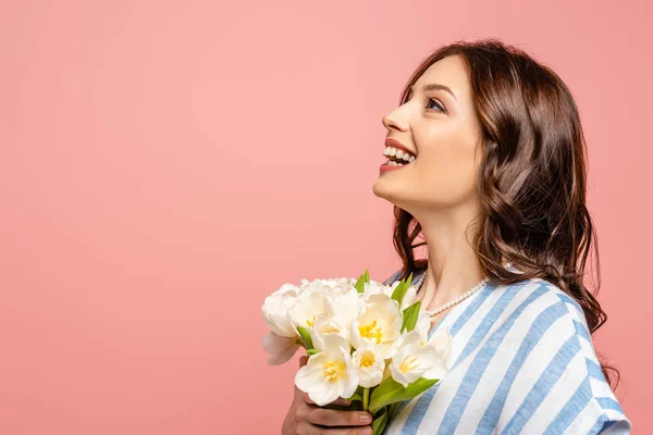 Chica feliz mirando hacia arriba mientras sostiene ramo de tulipanes blancos aislados en rosa - foto de stock