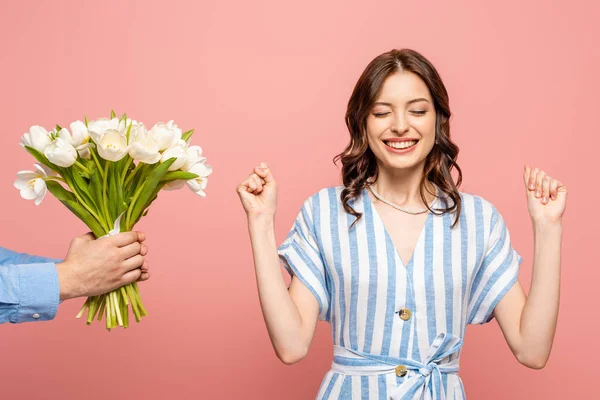 Vue recadrée de l'homme présentant bouquet de tulipes blanches à la jeune femme excitée montrant geste gagnant isolé sur rose — Photo de stock