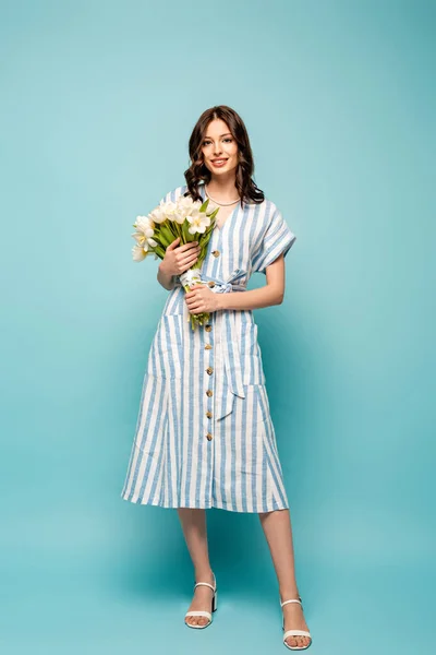 Vue pleine longueur de belle jeune femme tenant bouquet de tulipes blanches tout en souriant à la caméra sur fond bleu — Photo de stock