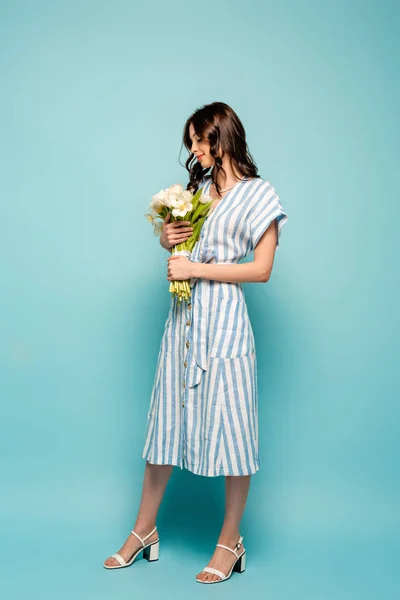 Volle Länge Ansicht der attraktiven jungen Frau hält Strauß weißer Tulpen auf blauem Hintergrund — Stockfoto