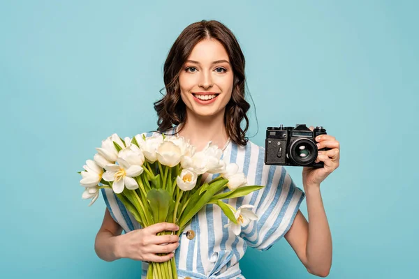 Happy young woman holding digital camera and bouquet of white tulips isolated on blue — Stock Photo