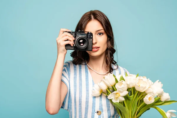 Atractiva joven mujer tomando fotos en cámara digital mientras sostiene ramo de tulipanes blancos aislados en azul - foto de stock