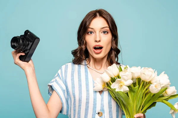 Surprised young woman holding digital camera and bouquet of white tulips isolated on blue — Stock Photo