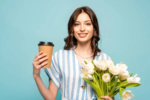 Feliz jovem segurando café para ir e buquê de tulipas brancas isoladas em azul — Fotografia de Stock